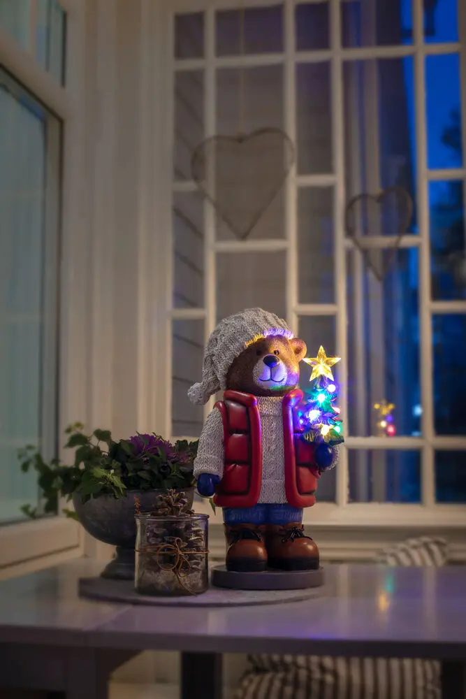 Kleine Teddybeer 38cm Veelkleurig Van Konstsmide - Er staat een felverlichte kerstboom en een lantaarn in, wat voor een gezellige sfeer zorgt.