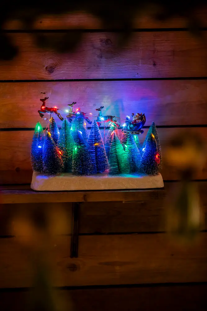 Kerstman Met Slee 22cm Veelkleurig Van Konstsmide - Kerstversiering met de Kerstman in een slee die over besneeuwde sparrenbomen vliegt.